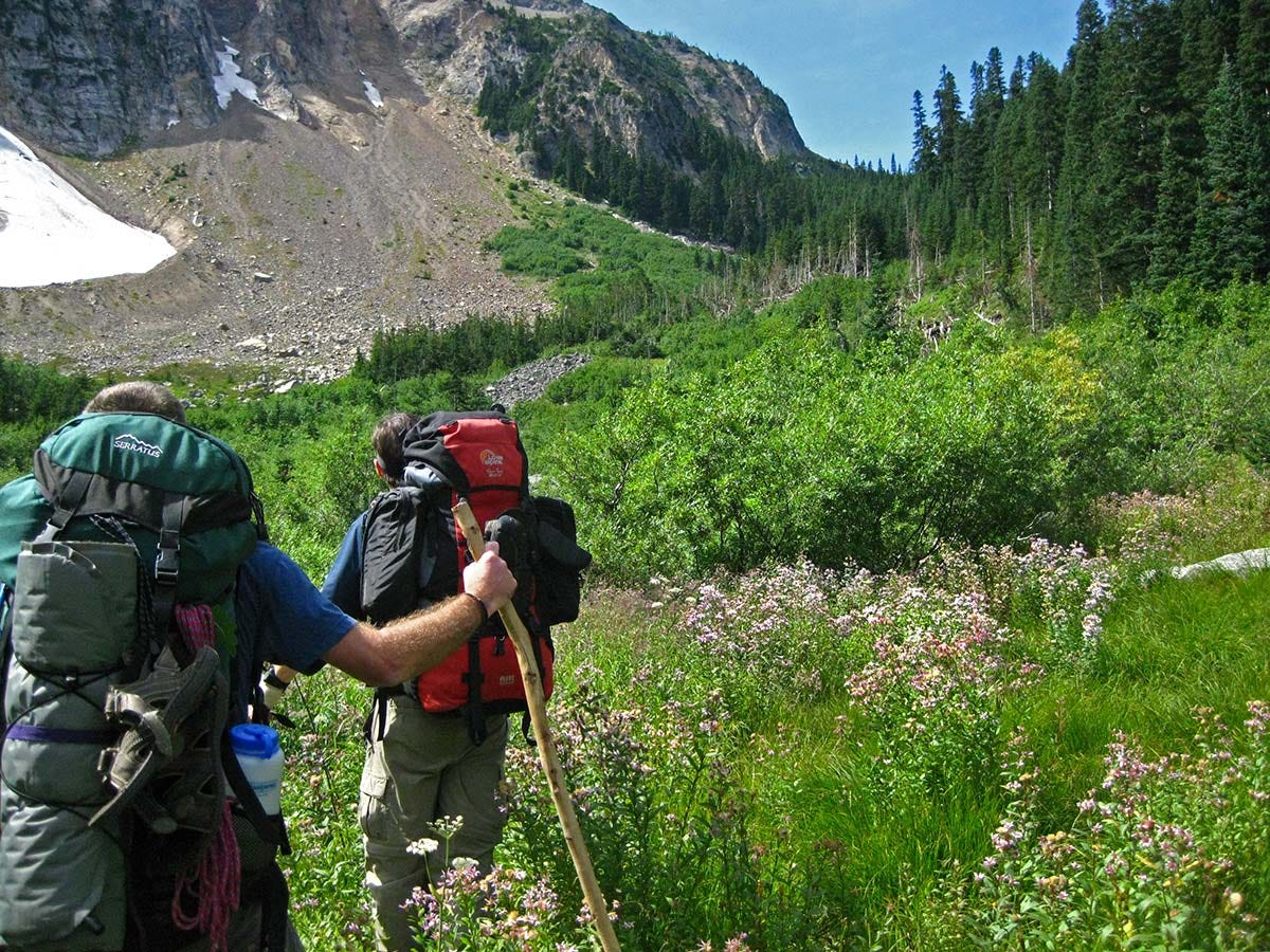 two men with loaded large packs, shoes and pads strapped to the outside, look up at a willow-filled valley beneath scree fields