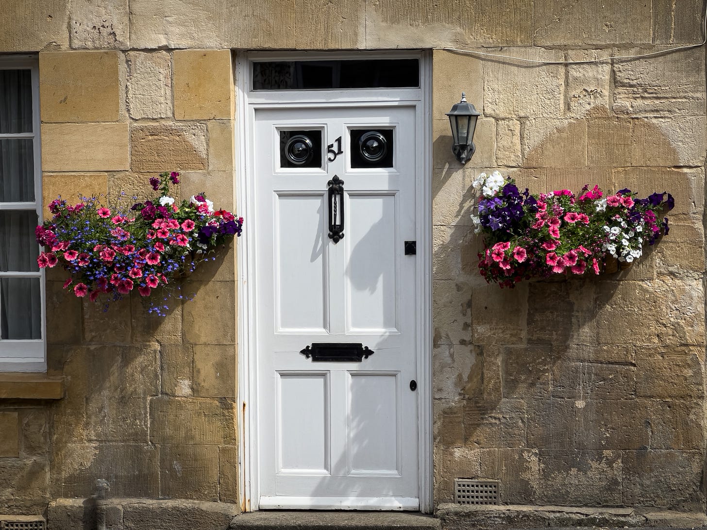 A white wooden fron door with two small windows near the top and the number 51 between the panes, a letter drop is centered on the lower third of the door. Pink, purple, and white petunias flank the door in floor pots adhered to the wall of the yellow-stoned house