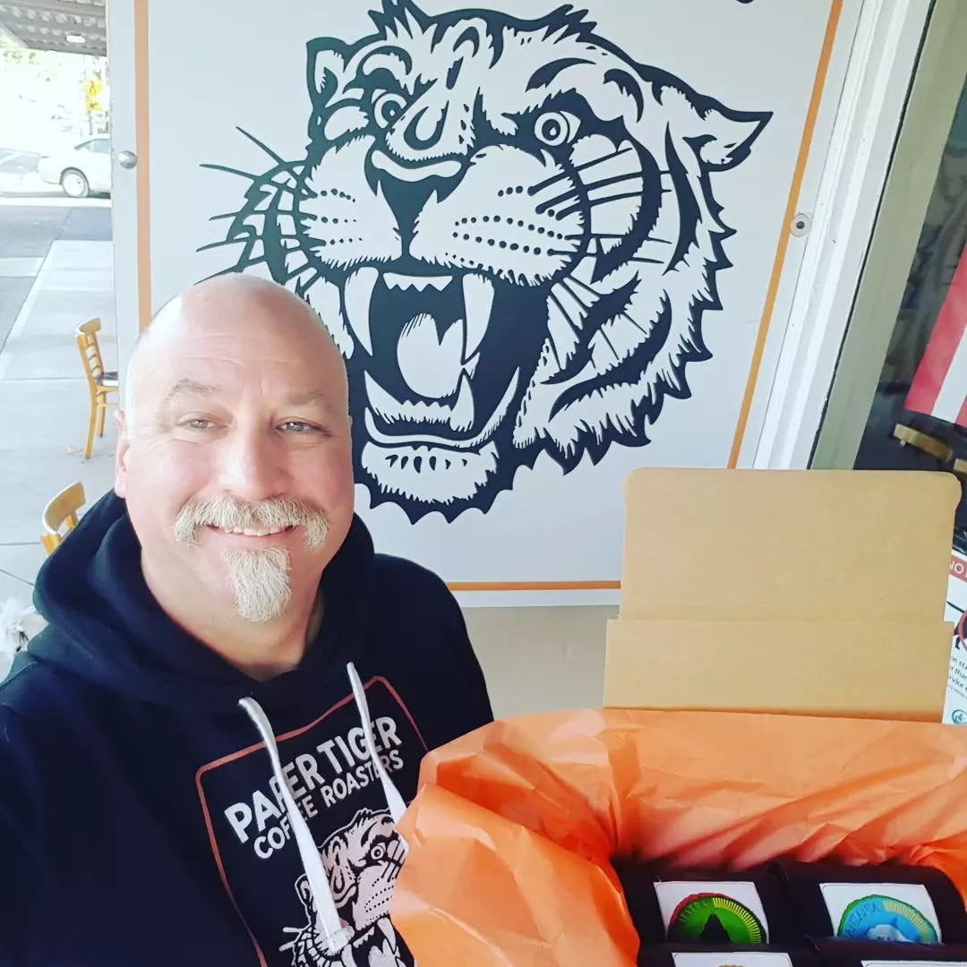 A bald man in a black hoodie sweatershirt stands in front of a sign with growling tiger holding a box of coffee beans.