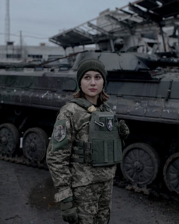 A woman in green camouflage stands in front of a burned military vehicle. 