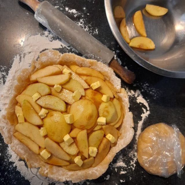 Image of a half-finished pie, the apple slices in the tin with dough to the side and a rolling pin.