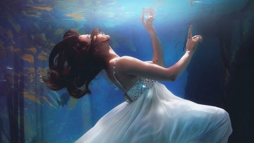 a woman in a white dress floats underwater, with her eyes closed and her face raised near the surface.