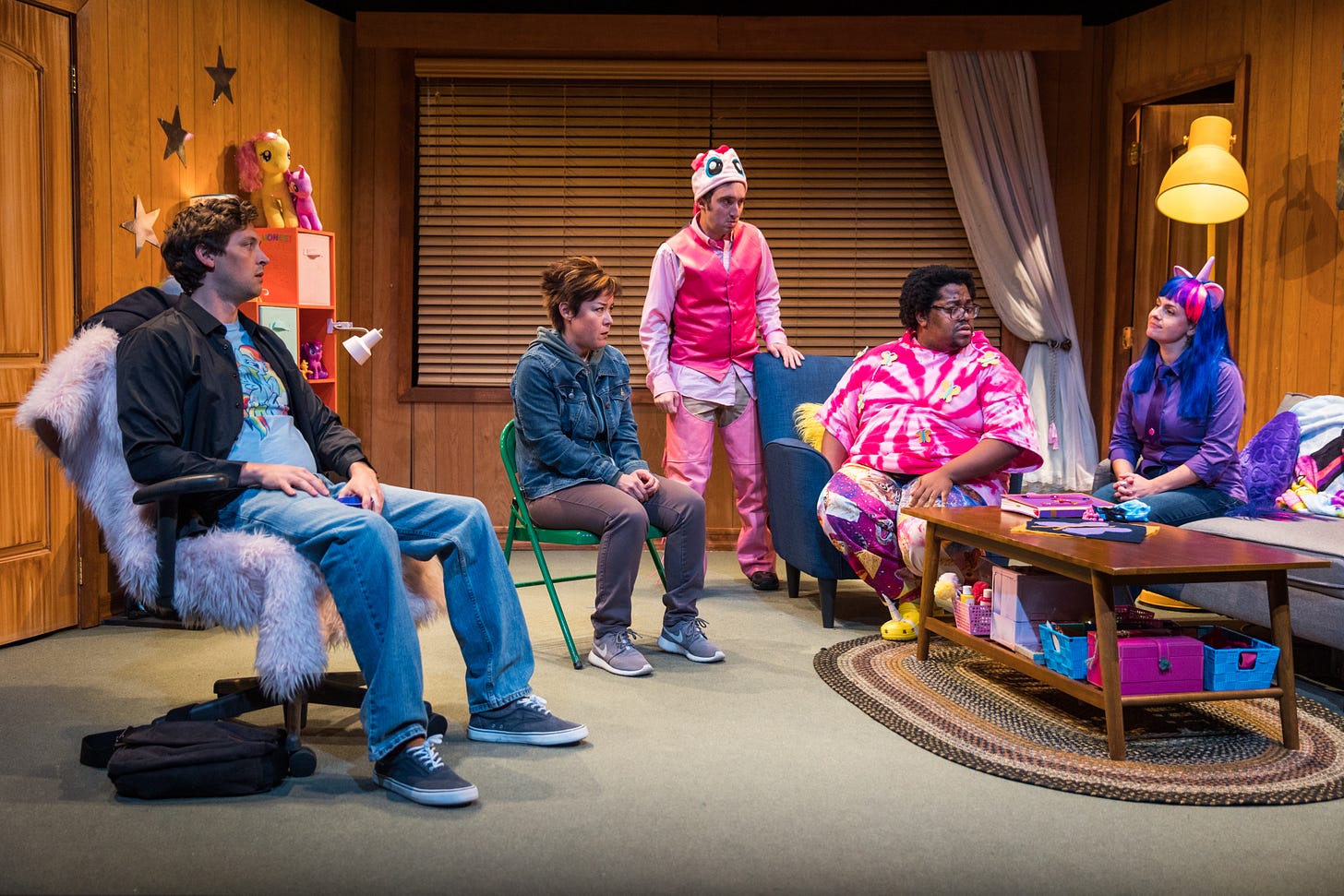 Five young people, three of which are wearing pony-themed costumes, sit in a circle in a small living room decorated with My Little Pony paraphernalia