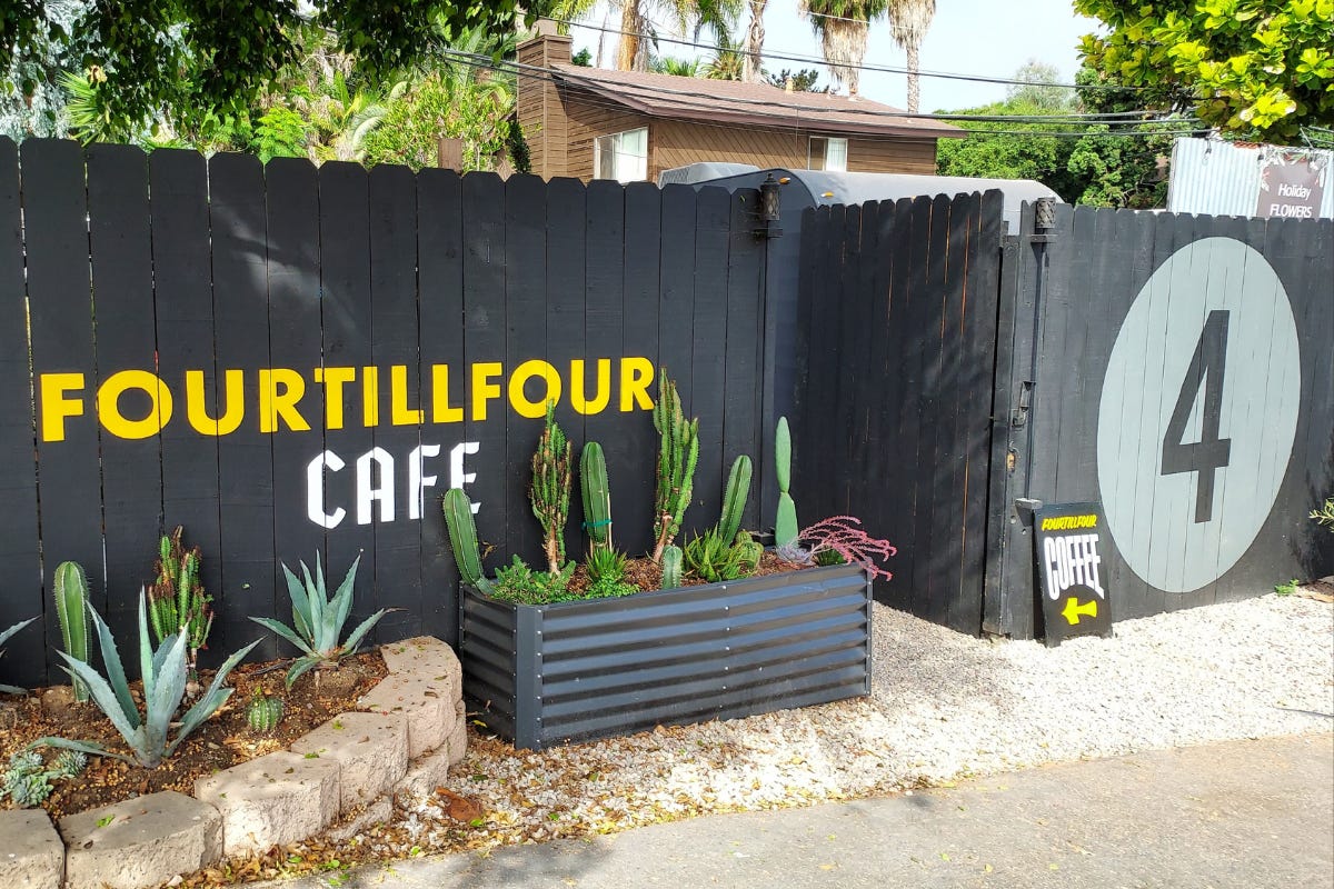A wood fence painted matte black. In yellow block text the cafe name "Fourtillfour" and in white, "Cafe." Two garden boxes, one of black metal roofing sheets cut to size and one of stone edging are filled with aloe and tall succulents. Splitting the fence is a gate.