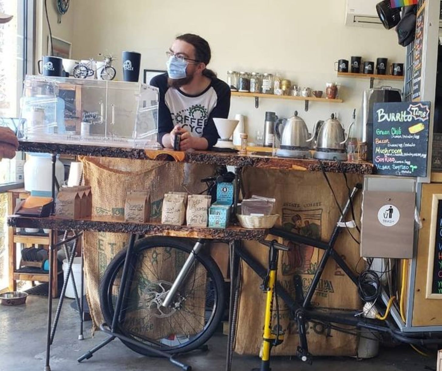 A white man with a ginger beard, glasses, ponytail and medical mask leans on a raw edge countertop at the coffee shop.