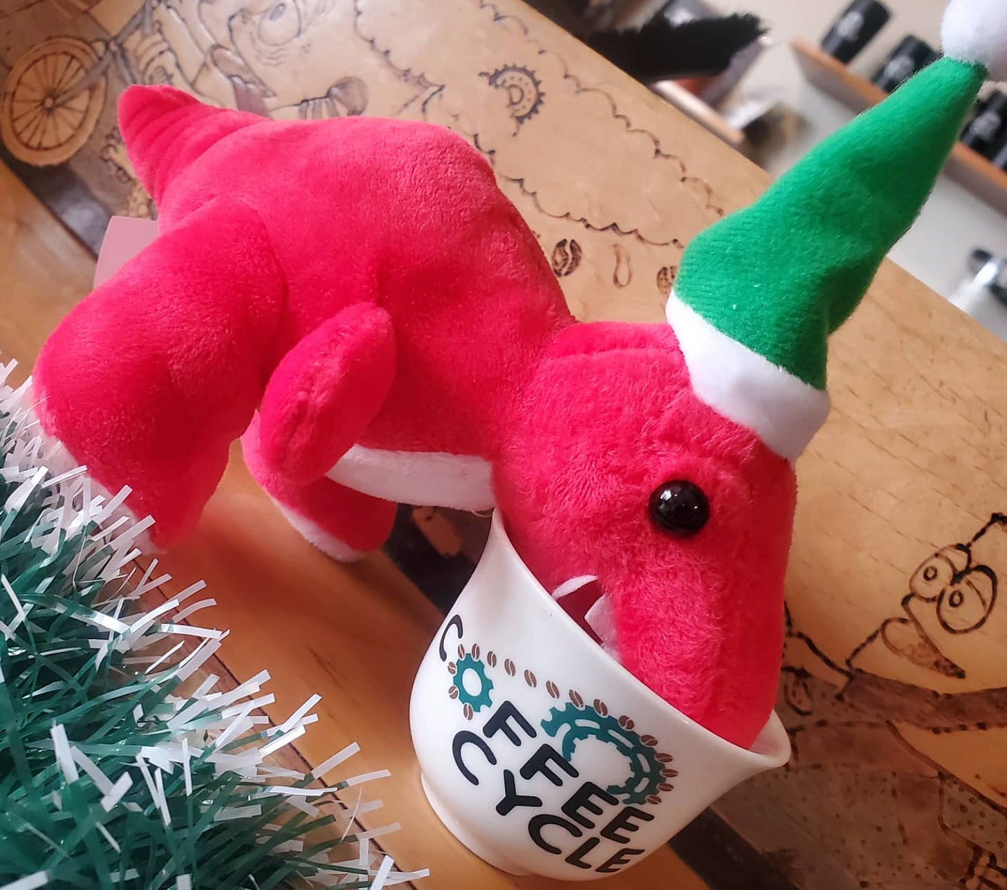 A red stuffed tyrannosaurus rex wearing a green elf hat drinking out of a demitasse espresso cup with the Coffee Cycle cafe logo on it. 