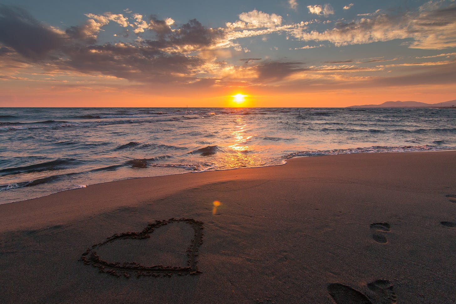The sun setting on a seashore with a heart drawn in the sand.