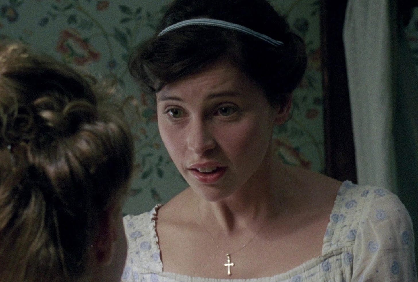 A close-up of Catherine Morland in a white Regency gown, a pale-blue Alice band, and a cross-chain around her neck, in earnest conversation. Felicity Jones portrays Catherine Morland. 