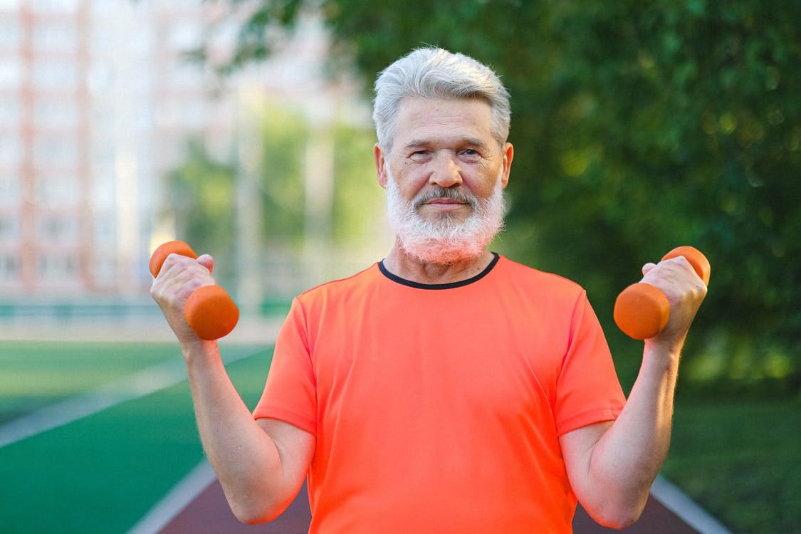 Smiling elderly man training with dumbbells on stadium and looking at camera in sunny day