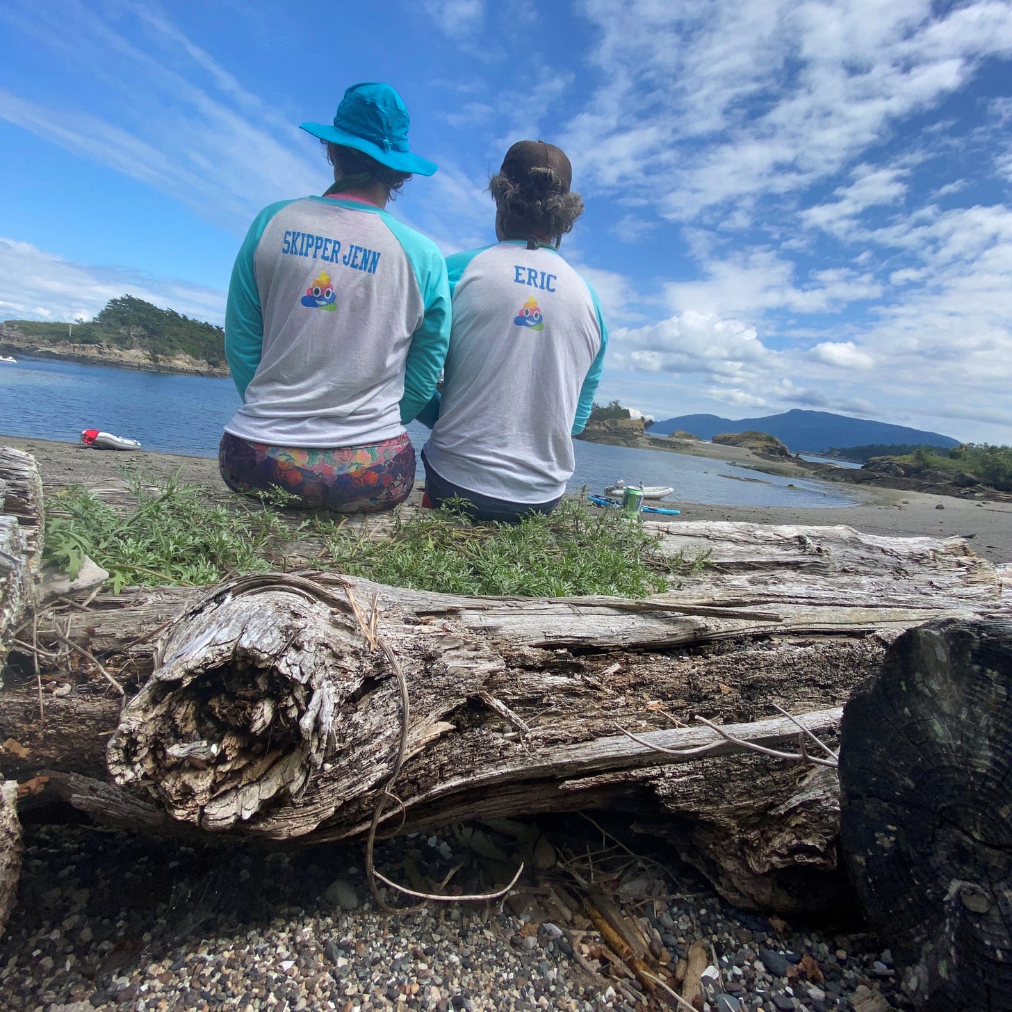 A couple sit on a log with their backs turned to us looking at the horizon. They wear matching team shirts that say Skipper Jenn and Eric on the back. 