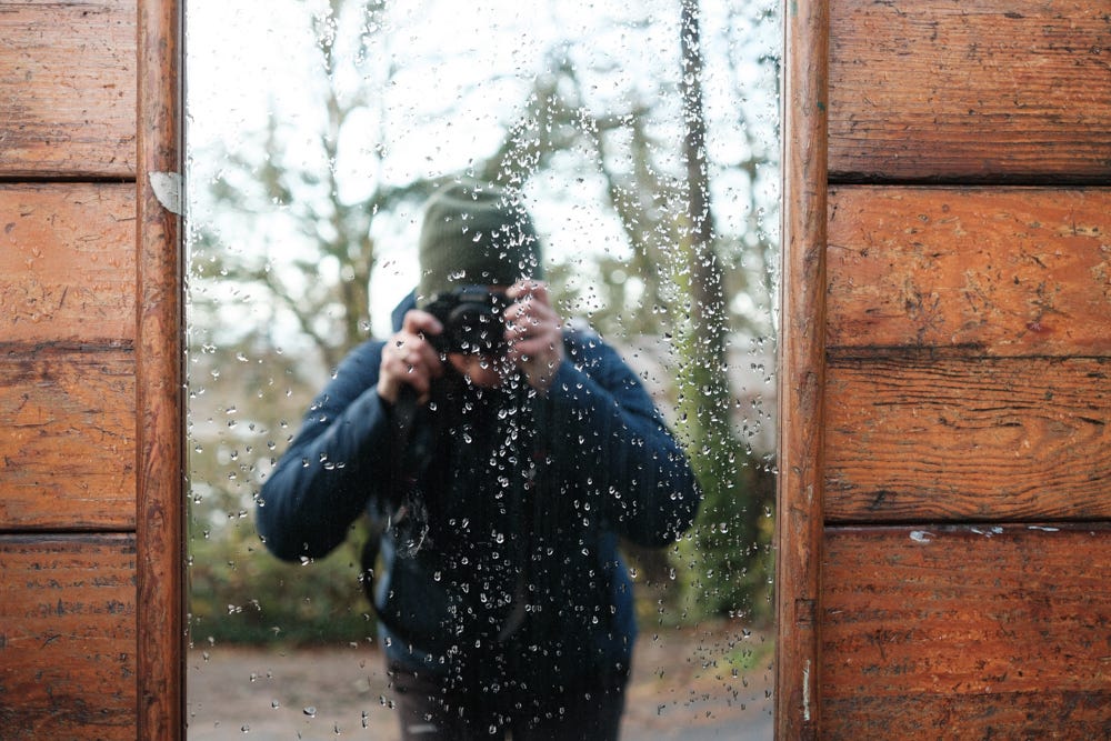 A woman in a blue winter coat and a green beanie takes a photo of herself with a camera in a mirror along a trail in Portland, Oreon.