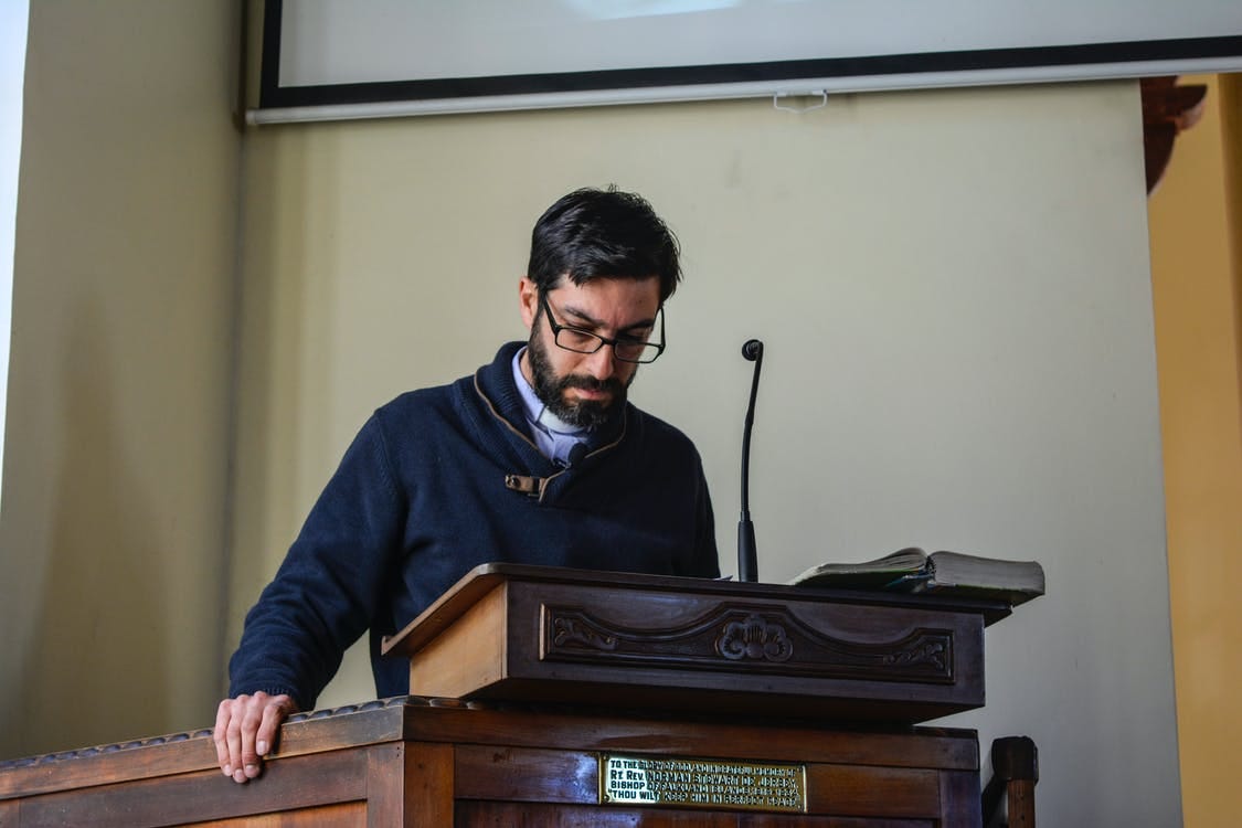 Serious bearded male teacher in casual clothes and eyeglasses reading lecture while standing at wooden podium in conference hall