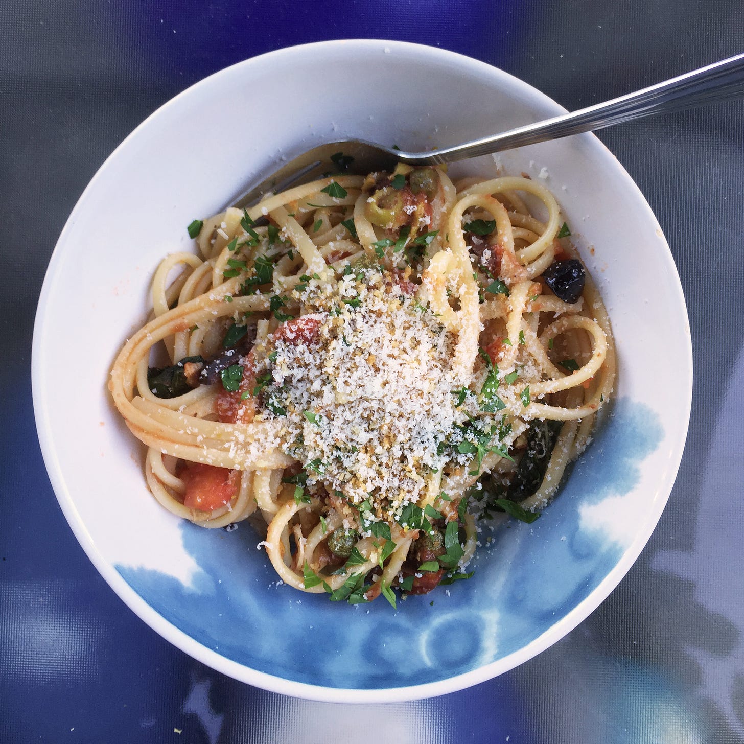 a white and blue bowl of linguine in puttanesca sauce, with parmesan and parsley on top.
