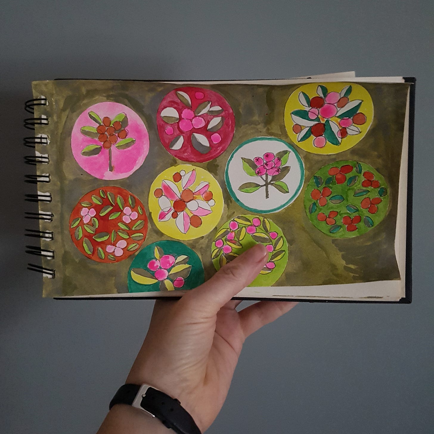 My hand holding up my sketchbook open at the page showing my leaf and berry designs on a murky green background