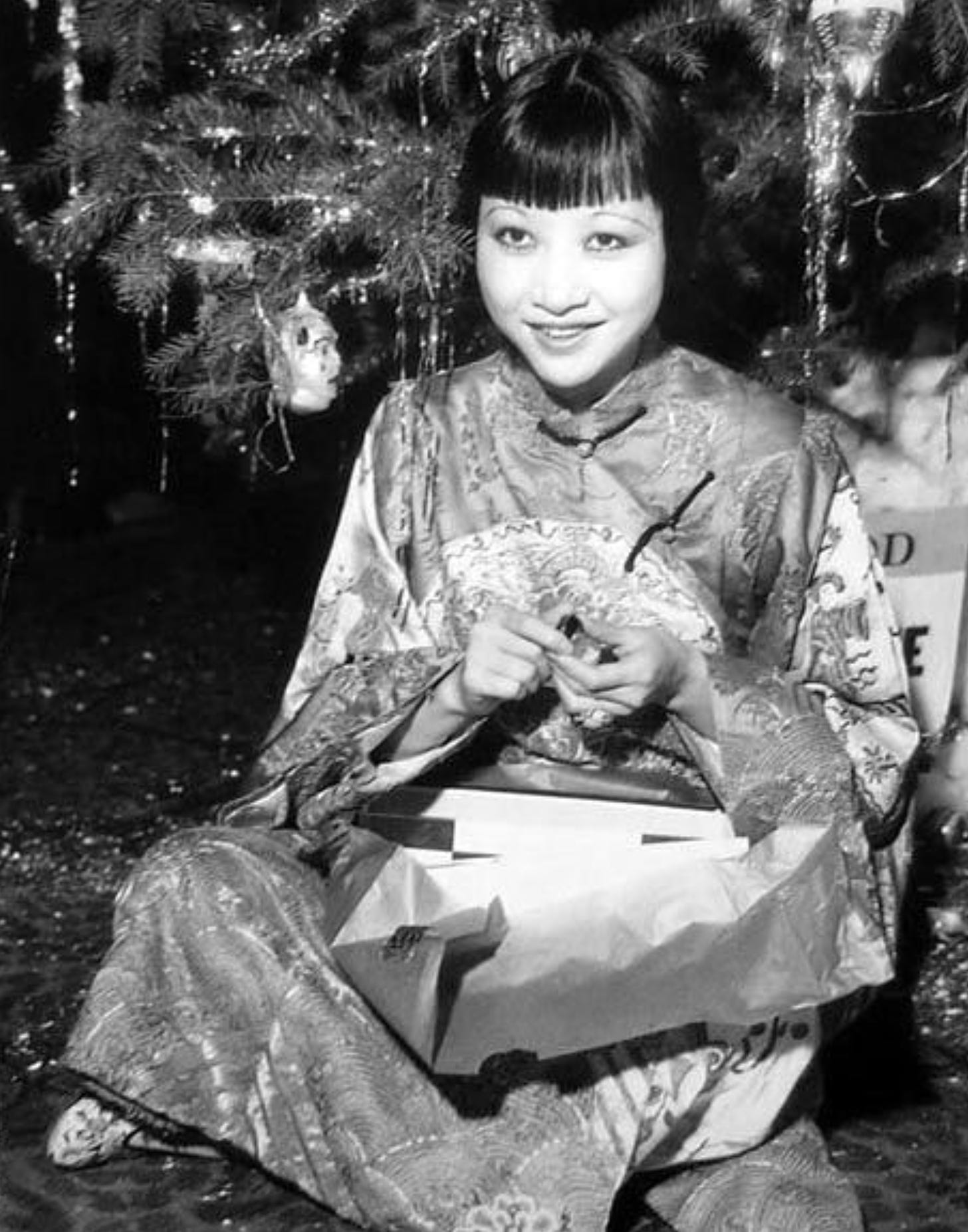 Black and white photo of Anna. May Wong opening a present underneath a Christmas tree