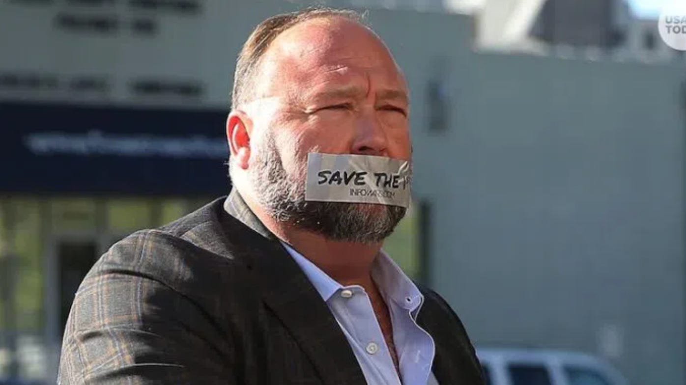 A picture of a middle aged white man with a greying short beard and short brown hair standing in the sun. He is wearing a dark plaid suit and a light shirt with the collar unbuttoned. He is wearing some type of grey tape over his mouth that has writing on it. The words "SAVE THE" is visible on the tape. Beneath that the words "INFOWARS" is visible.