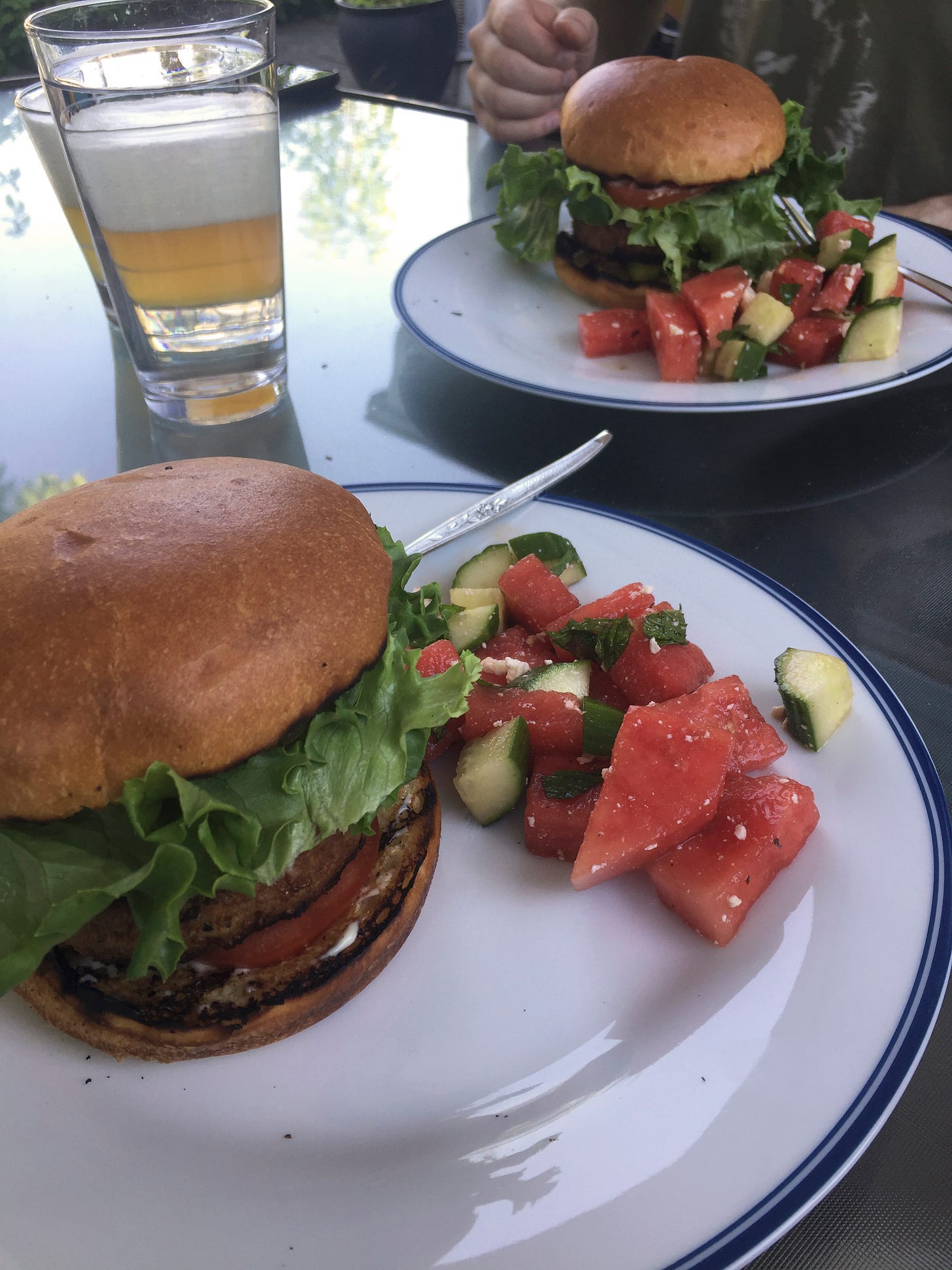 Two white plates with blue rims on an outdoor table. Each plate has a burger with a toasted bun and lettuce peeking out around the edges, next to a bright salad of watermelon and cucumber with mint and feta.