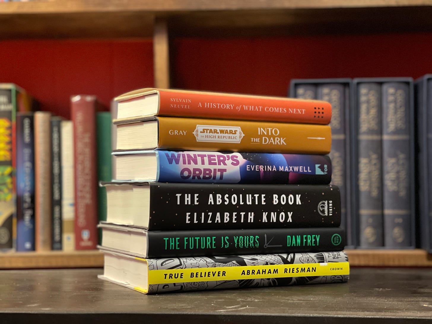 A colorful stack of books sits on a brown table, with a bookshelf in the background.