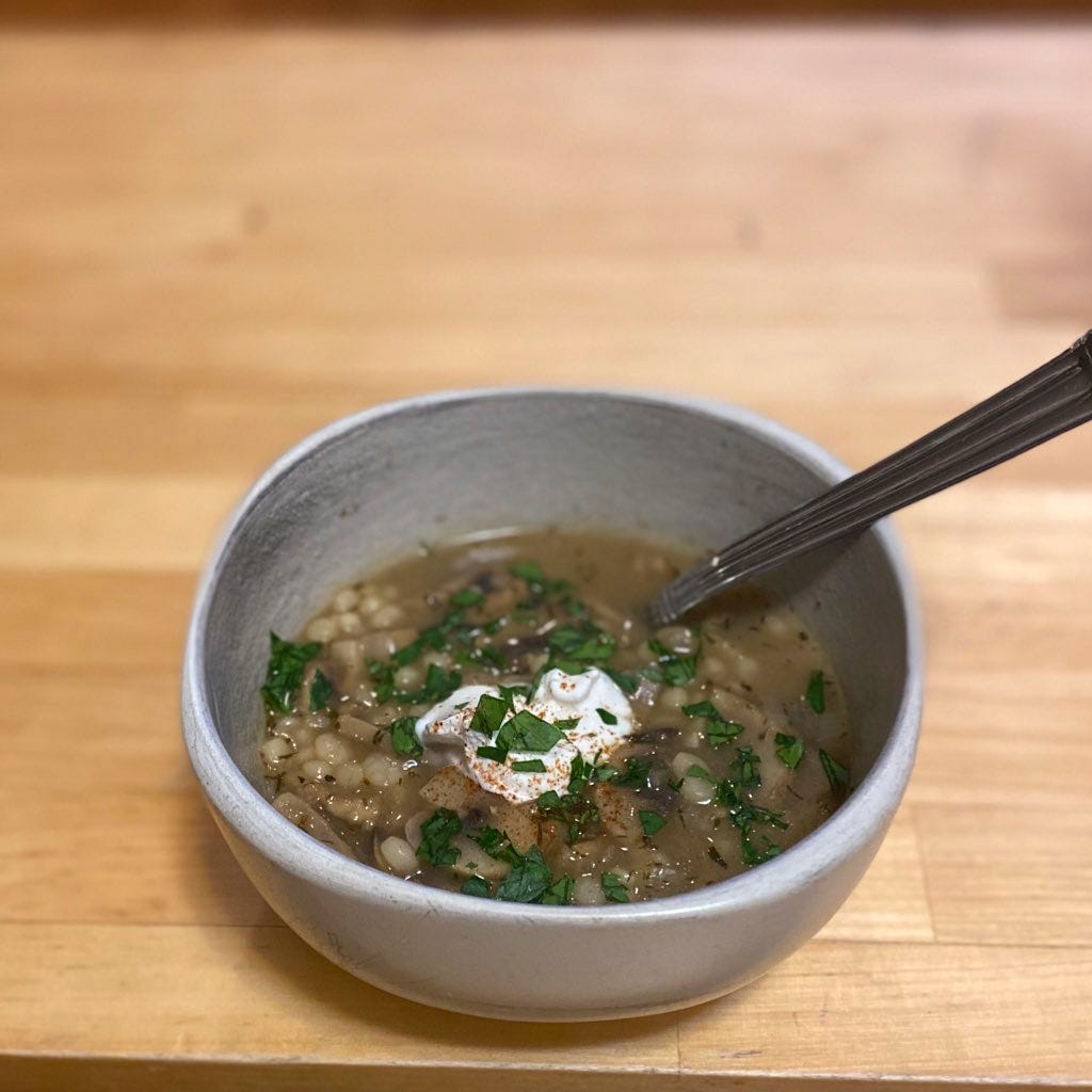 An off-white bowl of mushroom soup with pearls of couscous, and sour cream and parsley on top. A spoon sticks out at the right side.