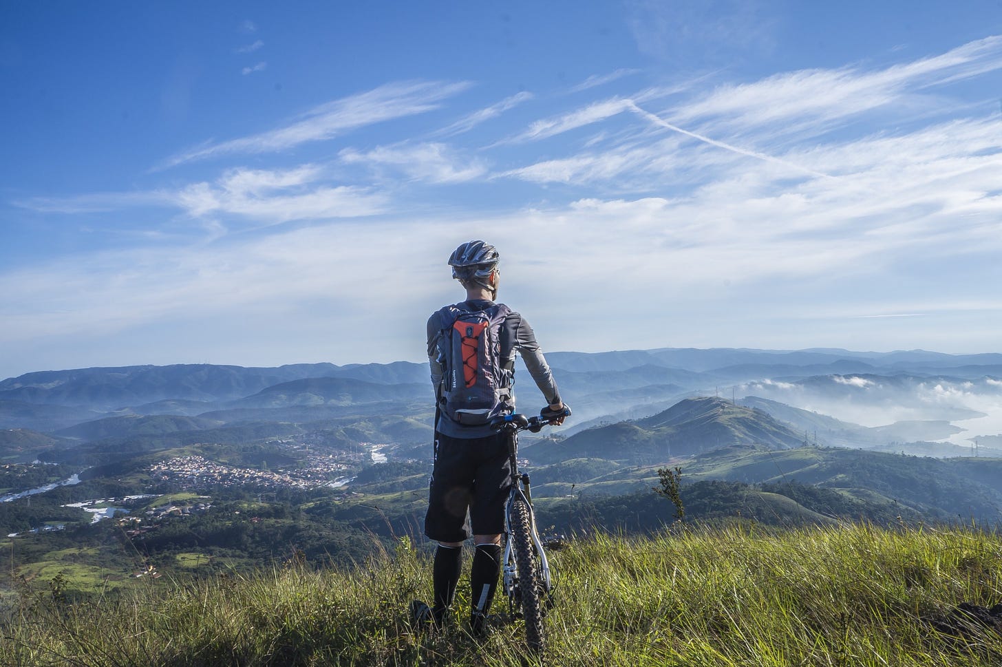 A cyclist off his cycle, standing on an expanse of grass and staring at the blue skies