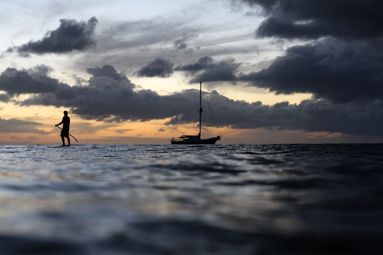 silhouetted individual on a sea board floating on dark waters in the sunset away from a distant sailboat.