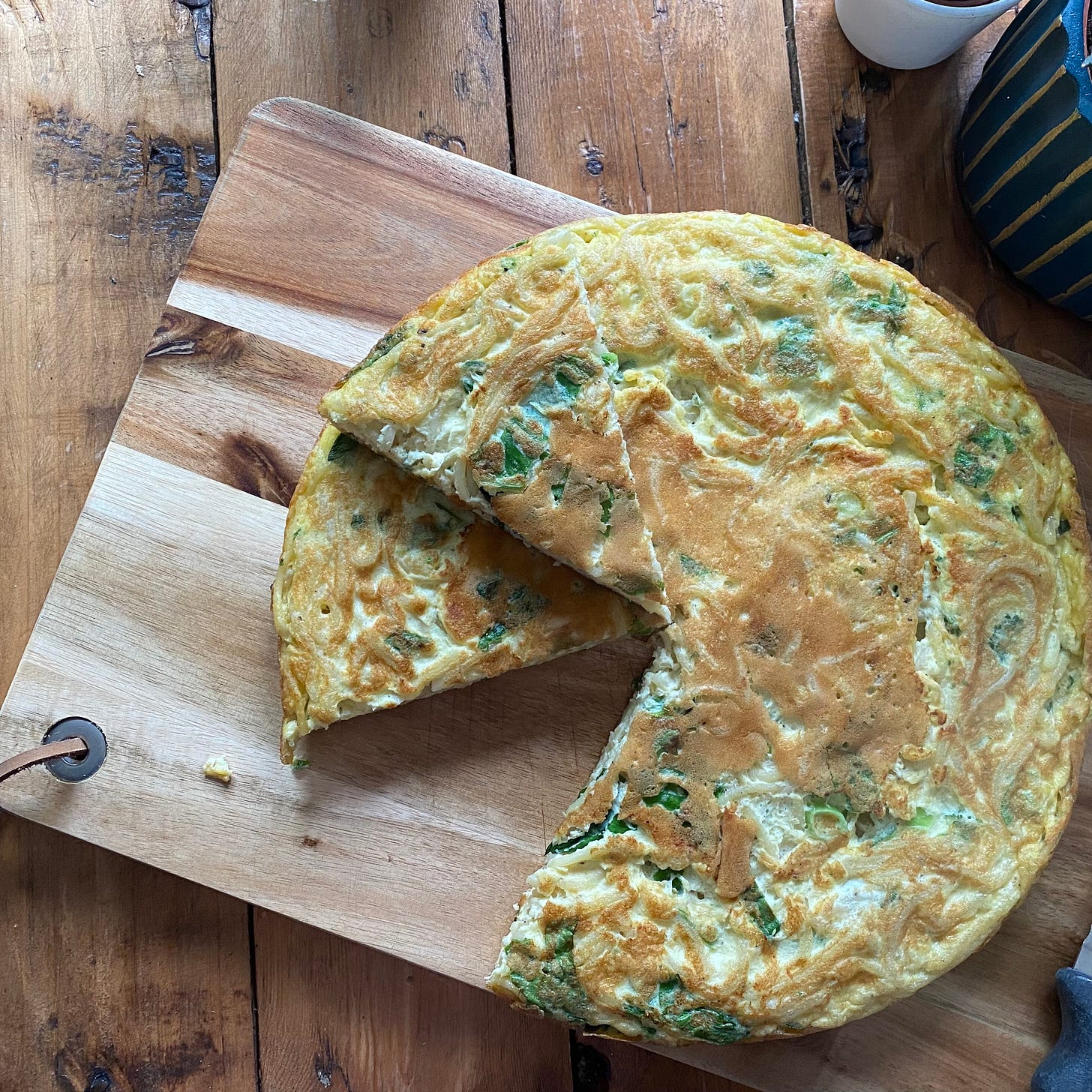 Round omelette served on a wooden board with a small wedge cut out. The frittata is made of cooked spaghetti, chard, onions, egg and cheese