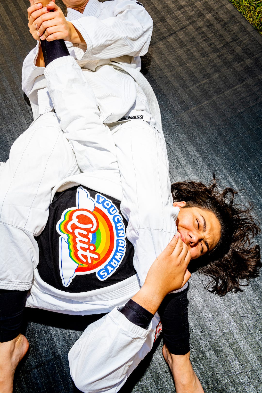 A martial artist in a submission hold, wearing a white gi over a You Can Always Quit t-shirt in black, lies on the floor a scrunches his eyes closed. He is just about to quit, and rightly so.