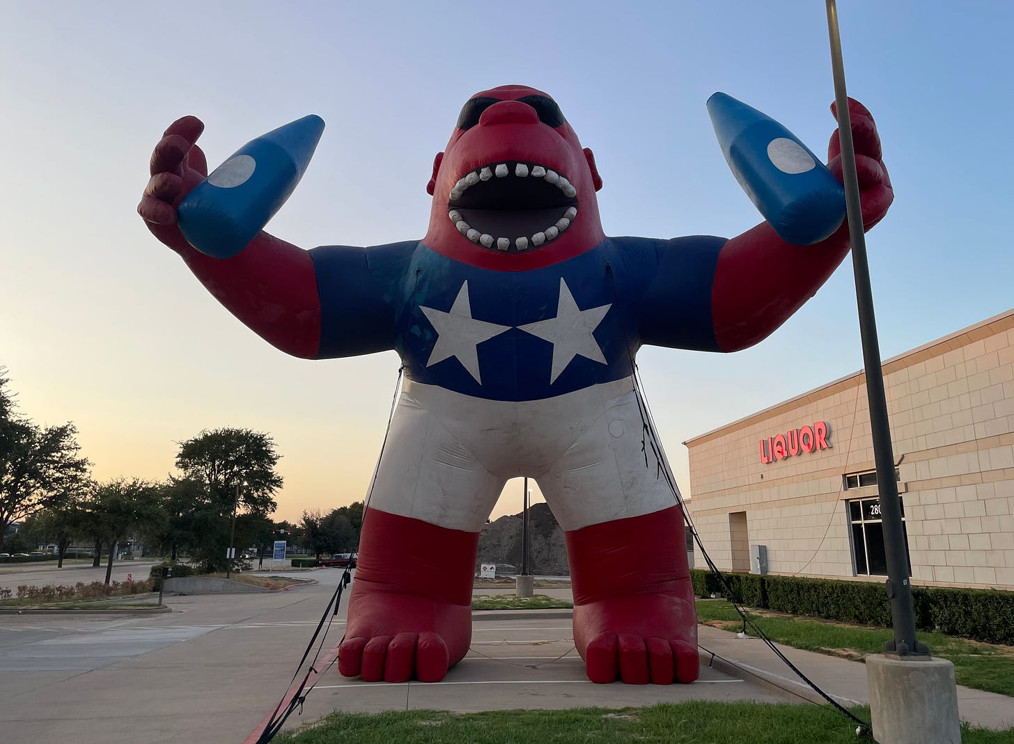 A red inflatable gorilla in a blue shirt and white shorts, holding two bottles in front of a liquor store