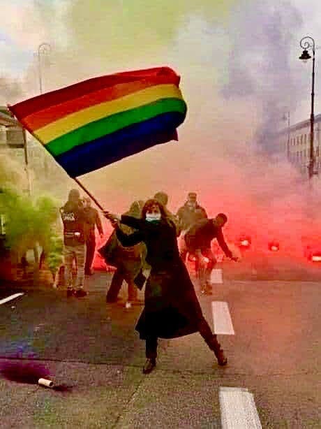 A woman waving a rainbow flag. She is wearing a face mask and is surrounded by tear gas in a protest.