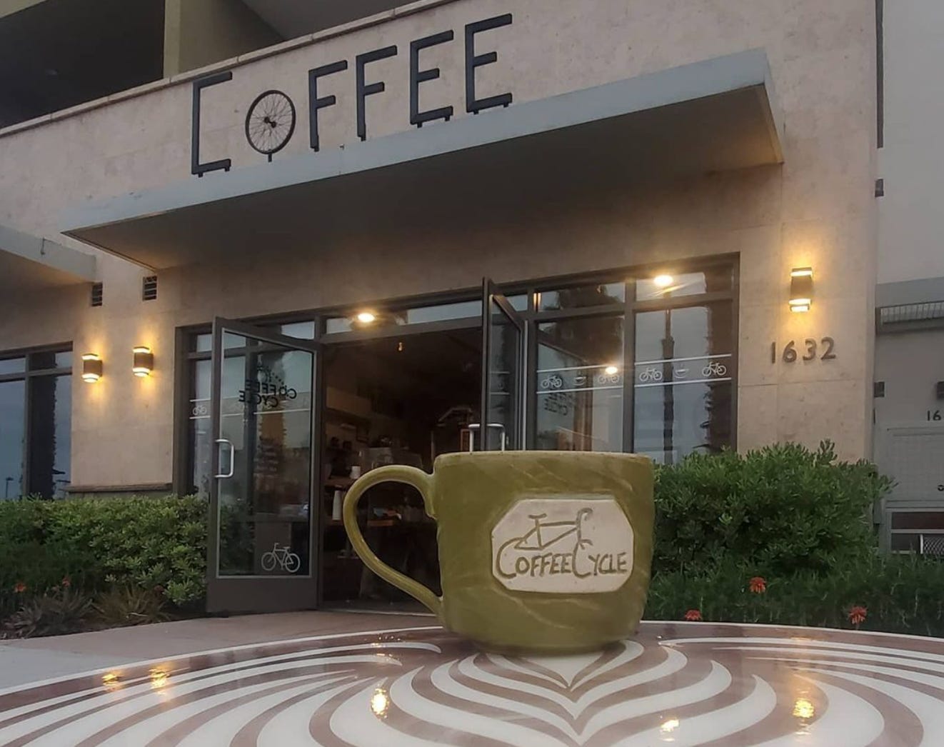 A close up of a green, hand made clay coffee mug with the Coffee Cycle logo in tan on the side. The mug is on a table with the entrance to Coffee Cycle in the background. The doors are open, and it says Coffee in big iron letters over the door.