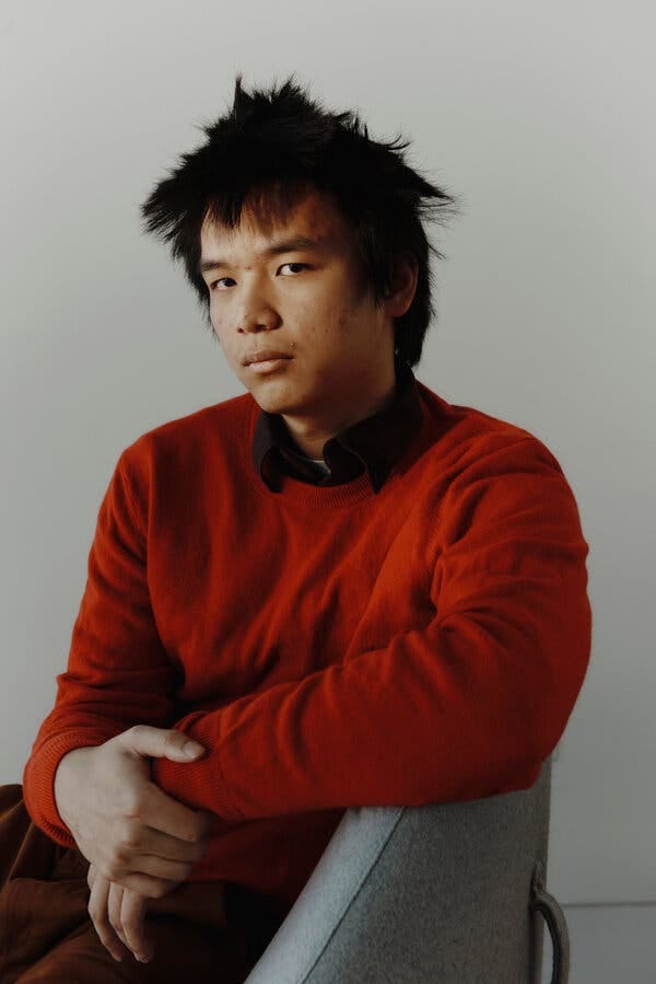 Max Li sits in a three-quarter pose, in front of a gray background. He has on a red sweater. He looks at the camera from the side, with a serious expression.