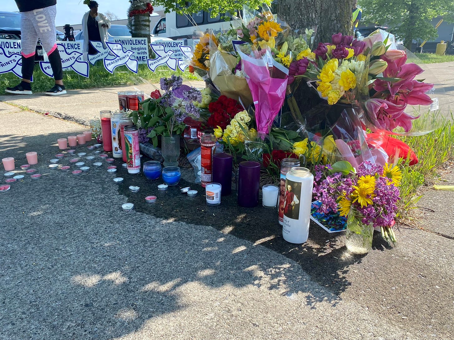 Flowers and prayer candles in vibrant yellow, pink, red, green and purples gathered under a tree beside signs saying "nonviolence" and "peaceprints"