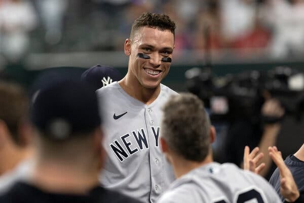 Wearing a gray Yankees road uniform, Aaron Judge smiles in the direction of the camera while standing on the dugout steps.