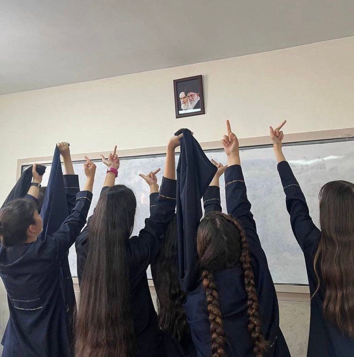 Schoolgirls photographed from the back raising their middle fingers and hijab at a picture of the ayatollahs who run their county