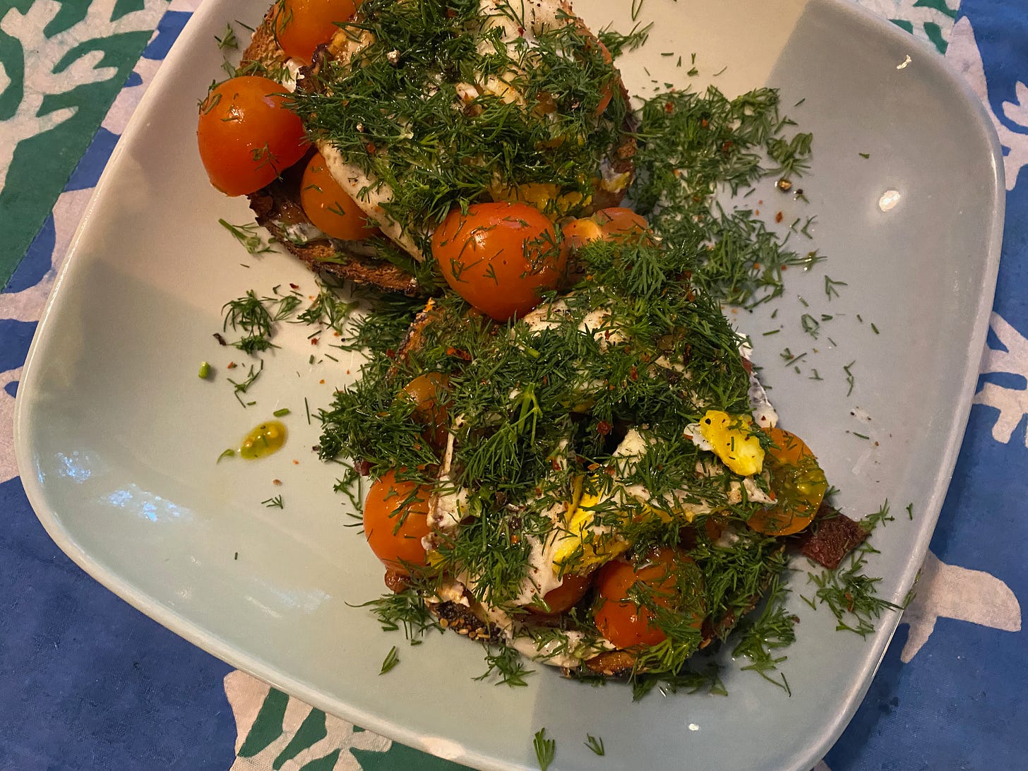 Two toasted bagel halves on a square ceramic plate. The bagels are piled with halved orange cherry tomatoes, a fried egg, and lots of chopped fresh dill.