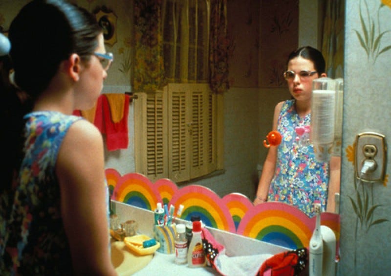 Preteen girl with dark ponytail, fair skin, and blue plastic glasses unhappily contemplating her reflection in the bathroom mirror, amidst happy rainbow decorations and eighties fixtures.
