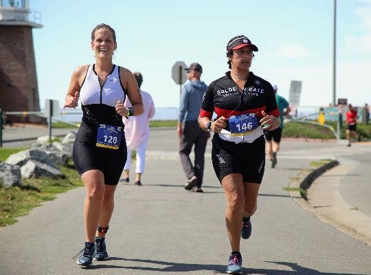 a mid-20s white woman smiling while running next to a 40s indian woman in a triathlon race