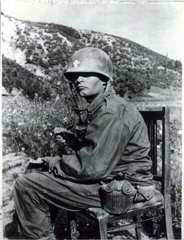 A man in combat dress looks out from under a helmet while writing a letter. The helmet has the insignia of a cross indicating that he is a clergyman.
