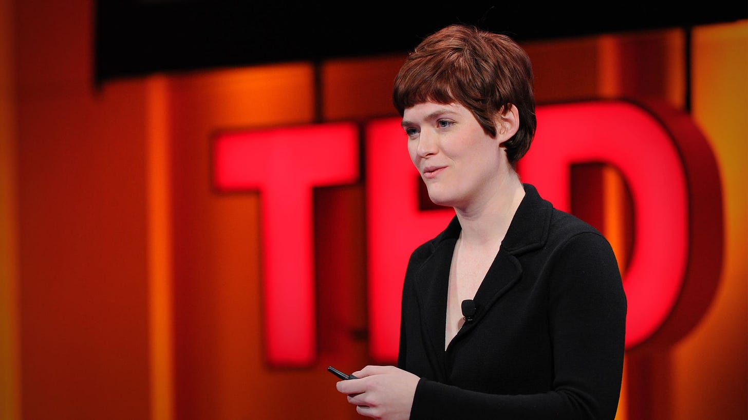 Foto de Amber Case com a logo do TED ao fundo. Ela tem cabelos ruivos e curtos, usa microfone de lapela e tem um passador de slides na mão.