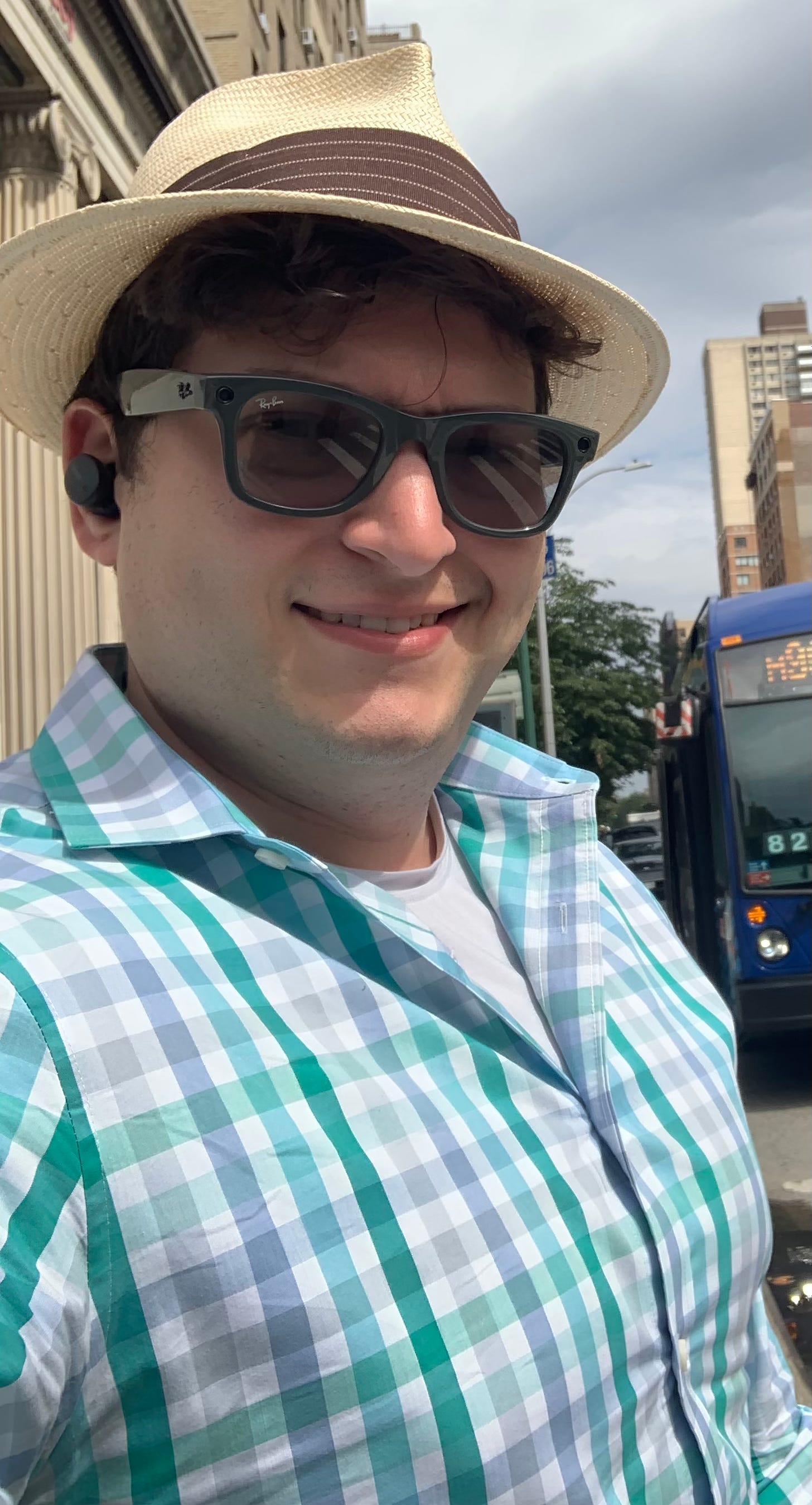 Image is me, a light-skinned, male-presenting individual, sort of smiling and sort of squinting out on a street in New York City. I am visible from the chest up, and I'm wearing a collared, button-down shirt with a plaid pattern that has crisscrossing lines of teal, blue, and grey. I am also wearing grey-framed Ray-Ban wayfarer sunglasses and a white panama hat with a brown band.