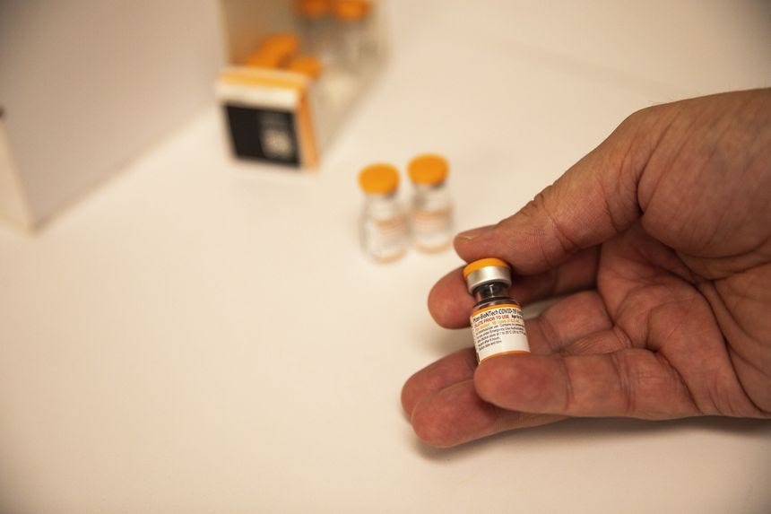 A hand holding a small vial of Pfizer-BioNTech's Covid-19 vaccine. More vials are displayed behind the hand.