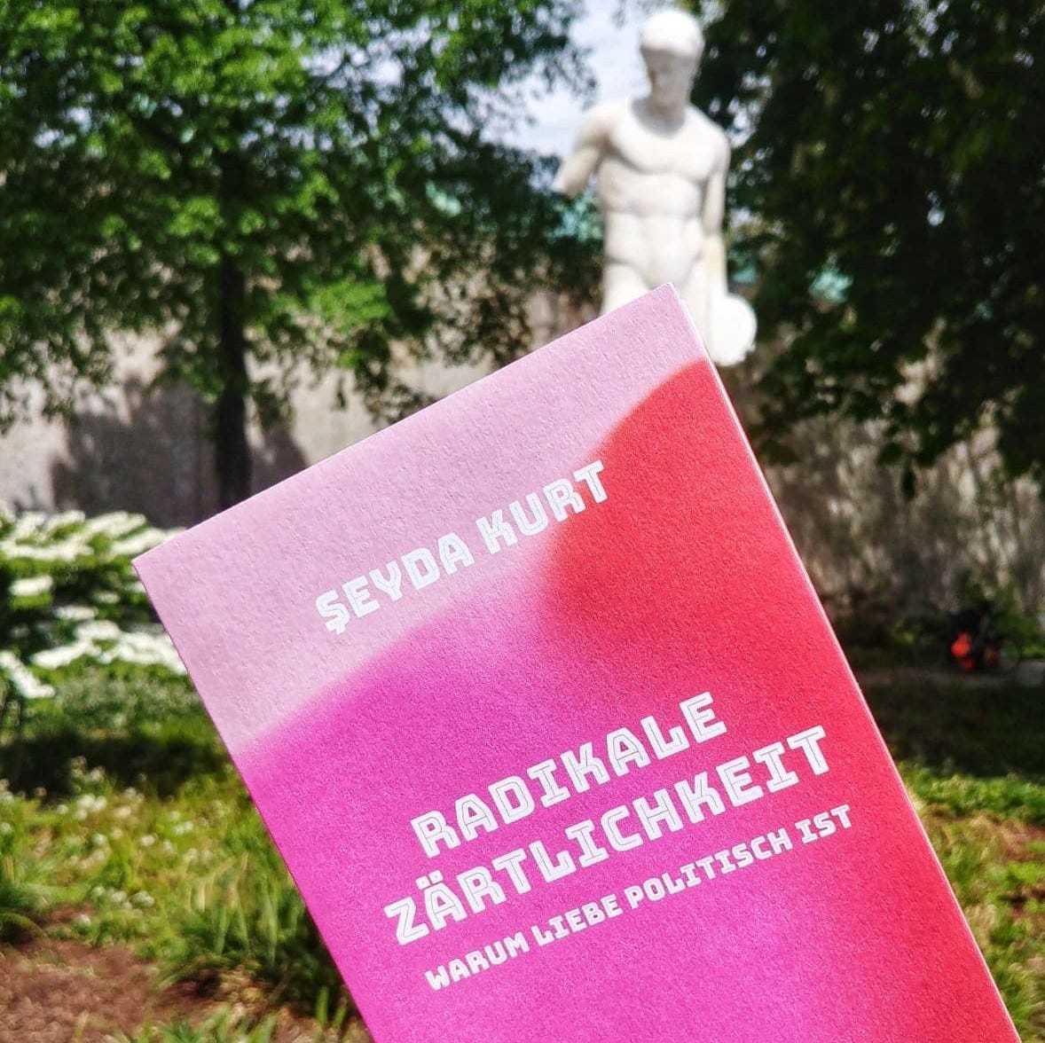 Titelbild: Das Cover des Buchs "Radikale Zärtlichkeit" von Şeyda Kurt wird so vor eine antike Männerkörper-Skulptur gehalten, dass eine Buchspitze den Penis verdeckt. Die Statue steht in einem sommerlichen Park.