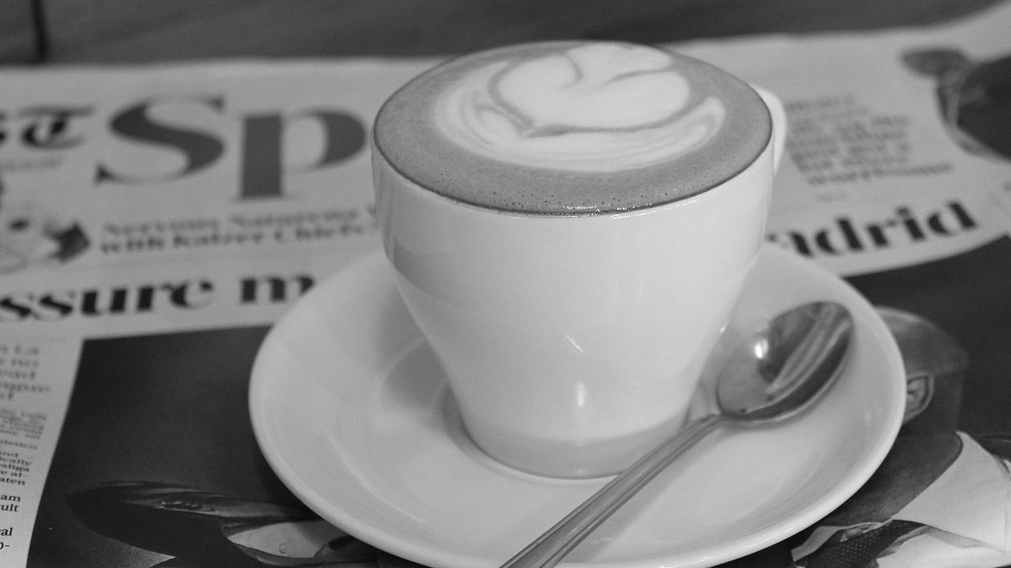 A white coffee cup with latte art and spoon sits atop a newspaper. Via Pixabay.