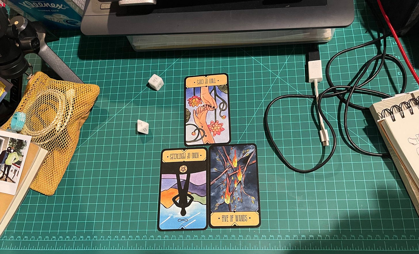 Picture of the tarot spread in the center of a mildly cluttered desk. Electronics, wires, and loose ephemera surround the three cards, Two of Wands, King of Pentacles, and Five of Wands.