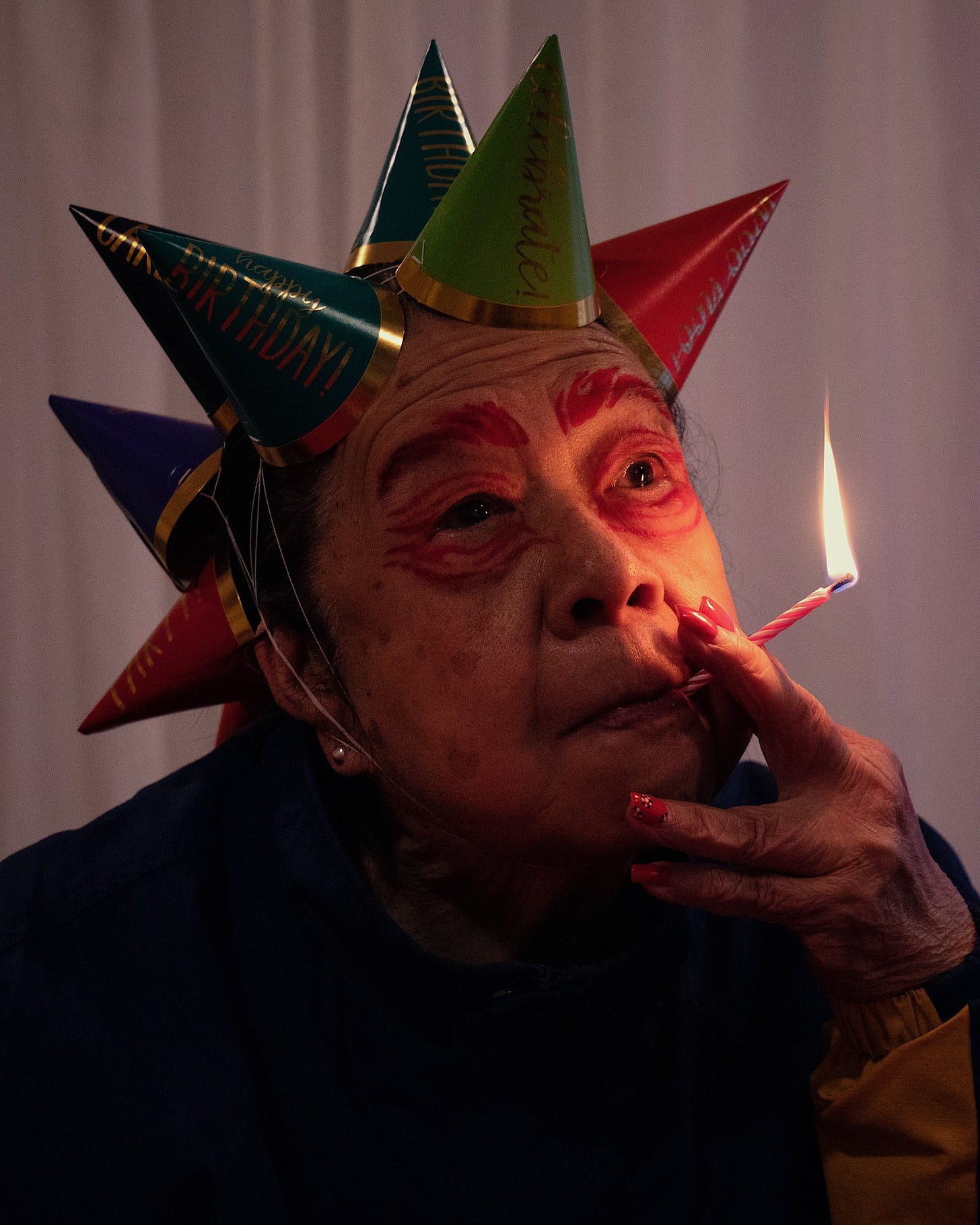 Fotografia de uma idosa com a cabeça coberta de pequenos chapéus cônicos de aniversário, fumando uma velinha de aniversário como um cigarro