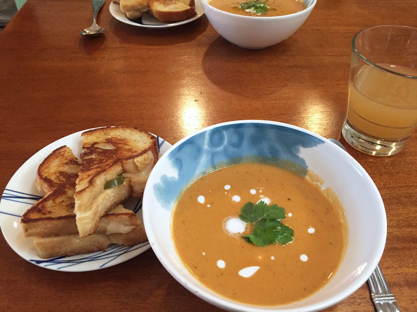 A bowl of orange tomato soup with dots of coconut milk and a few cilantro leaves. To its left, a small plate with two halves of a grilled cheese sandwich, jalapeño slices poking out.