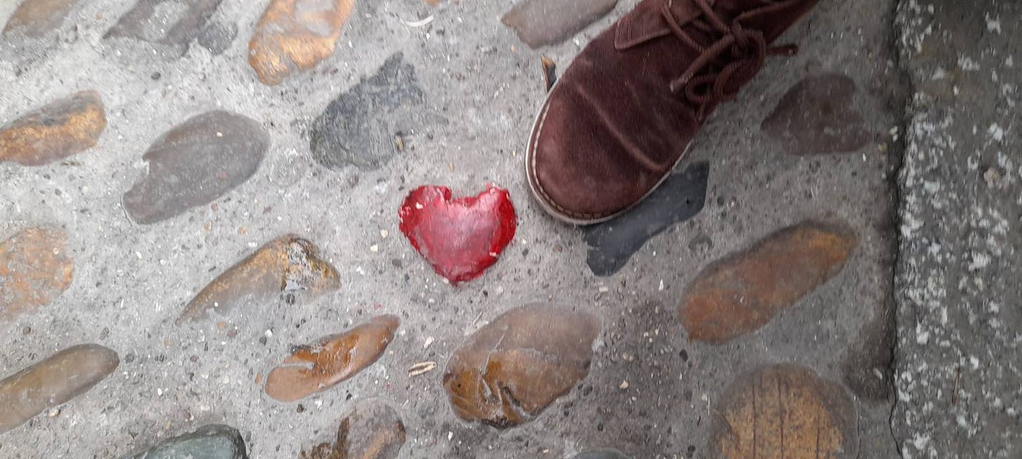 An image of a cobbled street. One of the stones is heart-shaped and painted red. A foot encased in a brown suede shoe is placed next to it.