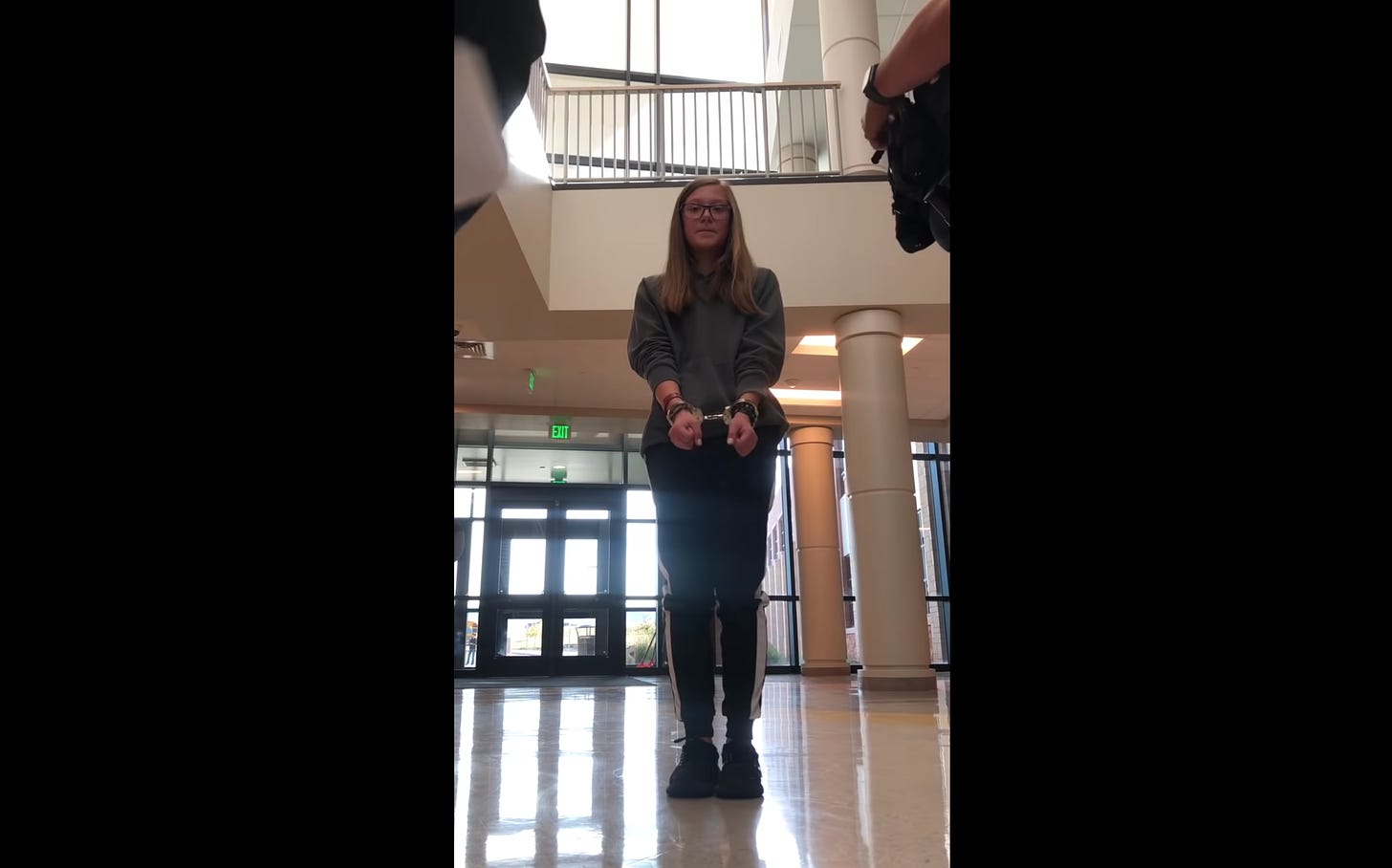 Handcuffed teenage girl stands in school atrium.
