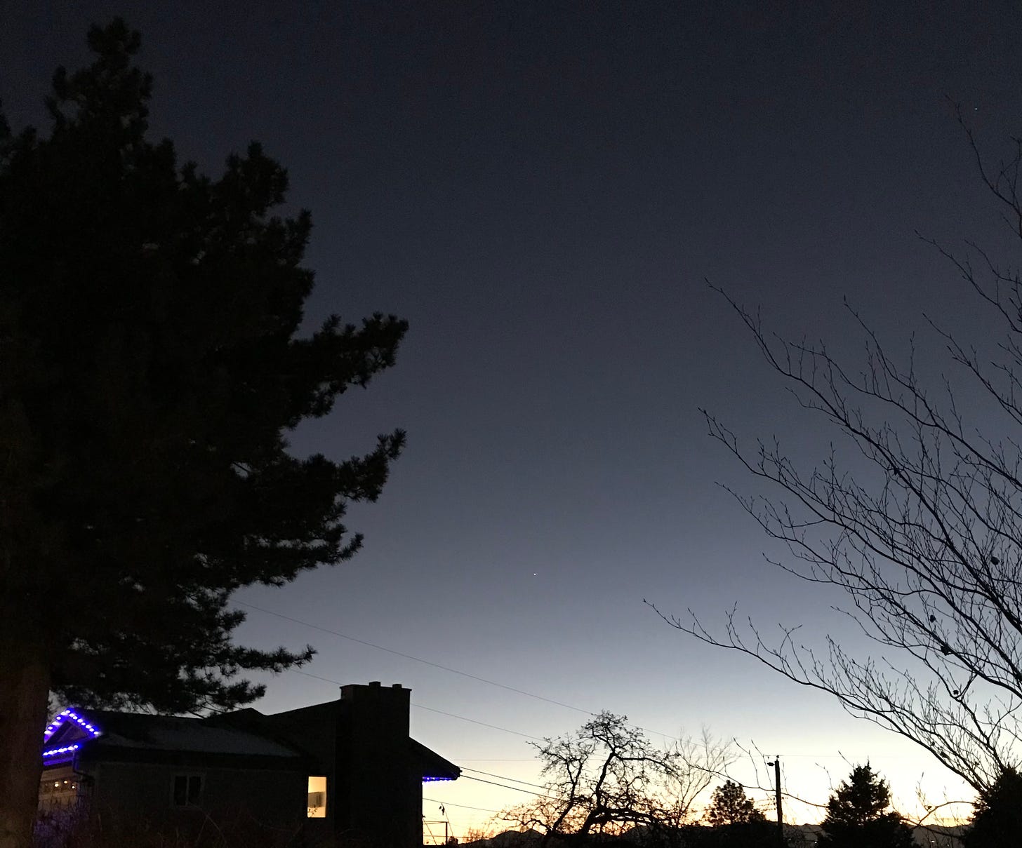 A view of the great conjunction of Saturn and Venus. The white dot of the two planets kissing is barely visible in the bottom third of the sky, which is a slate with gradient to yellow above mountains. In the foreground are a house with one lit window, a giant evergreen (black) on the left, and a branchy tree on the right.