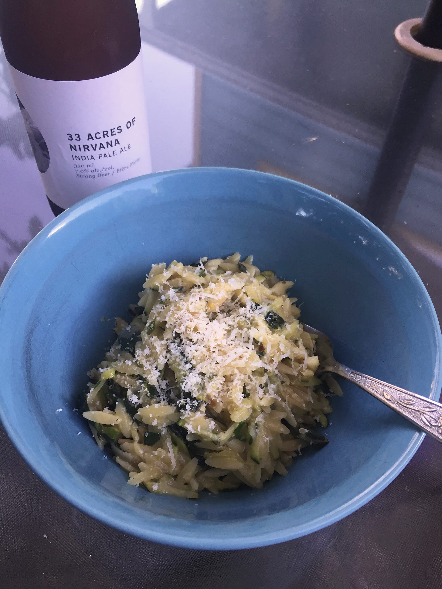 A blue bowl full of orzo, with shreds of pecorino on top and kale and zucchini visible throughout. Next to the bowl is a bottle of 33 Acres of Nirvana IPA.